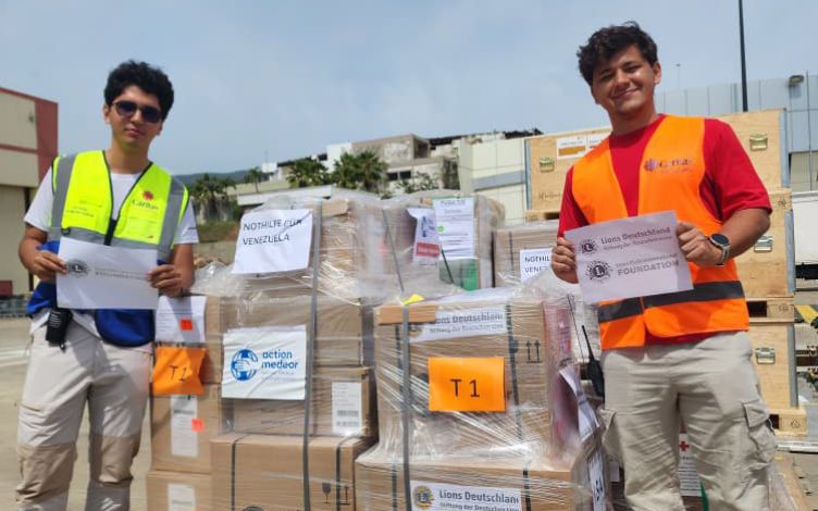 Two men wearing high-visibility vests stand in front of action medeor aid packages, holding signs reading “Lions Deutschland.” Photo: Joel de Abreu/Caritas La Guaira  